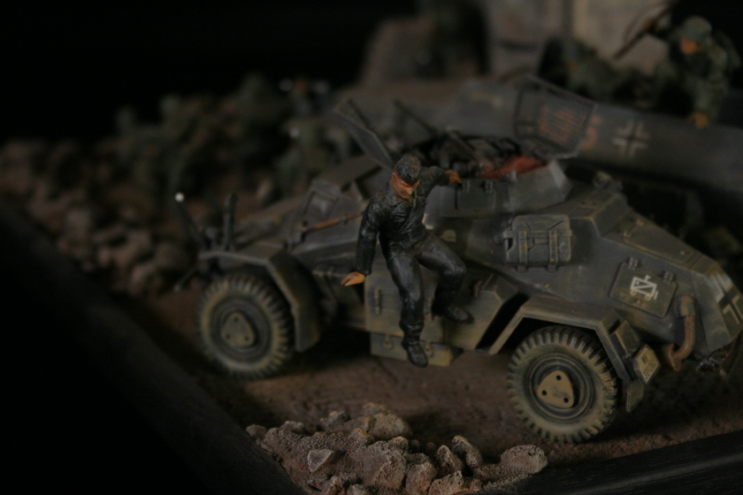 Close up view of a figurine of a man in an army uniform hanging off the side of an armored military vehicle driving in a rocky landscape. 