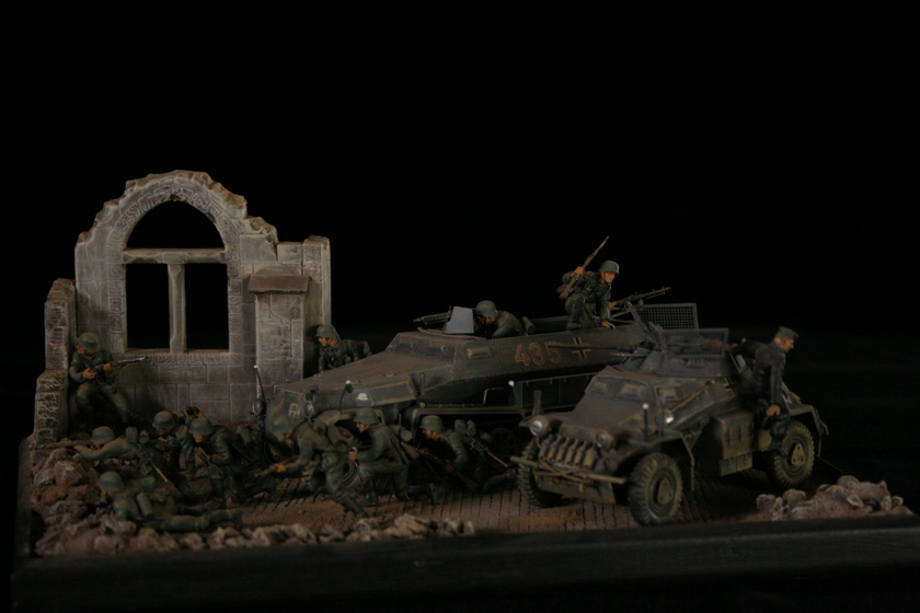 A model of two armored military vehicles sitting in a building, behind a group of men wearing military army uniforms crouched behind a crumbled wall. 