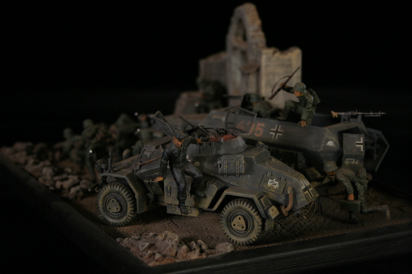 A model of two armored military vehicles sitting in a building, behind a group of men wearing military army uniforms crouched behind a crumbled wall. 
