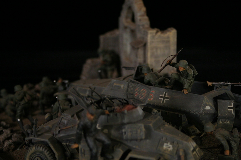 Close up view of a model of two armored military vehicles sitting in a building, behind a group of men wearing military army uniforms crouched behind a crumbled wall. 