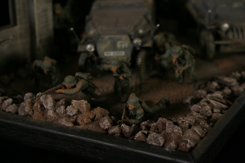 Close up view of a model of men wearing military army uniforms crouched behind a crumbled wall. Two armoured military vehicles are seen behind. 