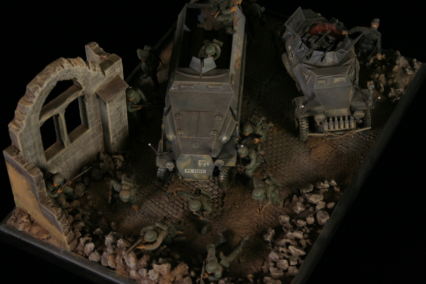 Overhead view of a model of two armored military vehicles sitting in a building, behind a group of men wearing military army uniforms crouched behind a crumbled wall. 