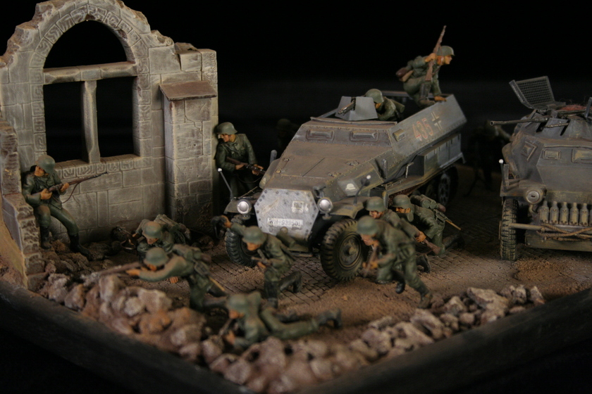 Close up view of a model of two armored military vehicles sitting in a building, behind a group of men wearing military army uniforms crouched behind a crumbled wall. 
