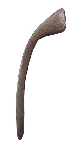 Wooden club with a thin handle made from brown timber, which bends and curves up to the right to a widened area on one end. 