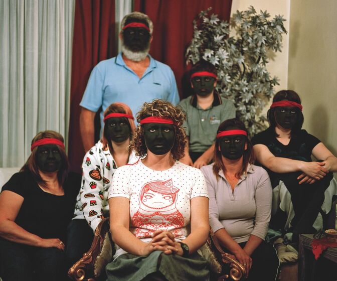 Photographic artwork of a group of seven men and women seated in a lounge room in front of a tree. Each person looks directly at the camera, has a red headband across their forehead, and has a face painted black.