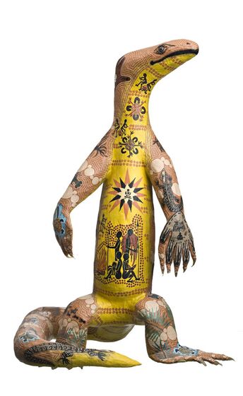 Sculpture of a brown goanna with a yellow belly, standing upright. across the surface of the sculpture are painted line and dot designs, including a multi-pointed star on the chest in white, brown and black, below which are four human figures. 