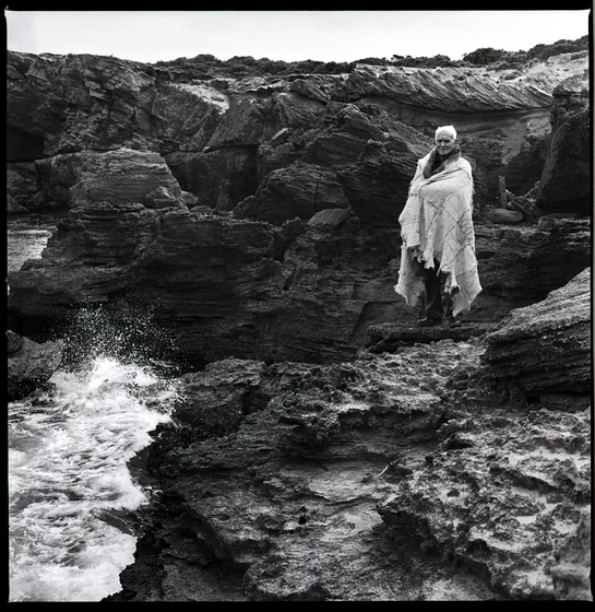 Elderly man holding a possum skin cloak around his shoulders, standing on beach-side rock formation, with ocean waves hitting the left side of the rocks.