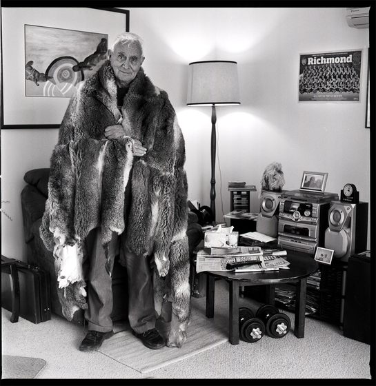 Elderly man wearing a possum skin cloak over his shoulders stands in a lounge room. Behind is an artwork of Indigenous Australian design. To his left is a Richmond Football Team club poster, a coffee table, lamp, and stereo system.