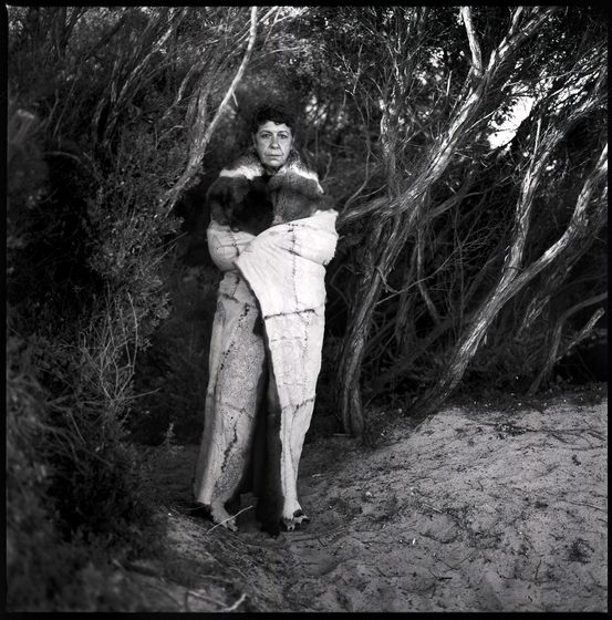 Black and white photograph of a woman with a possum skin cloak wrapped around her, standing on a sandy surface with shrub-like trees behind her.  