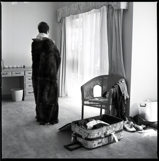 Black and white photograph of a woman standing in a lounge room, with a possum skin cloak wrapped around her shoulders, her back facing the camera. To her right is a wicker chair with clothes hanging off the arm, and an open suitcase with some clothes inside on the floor.