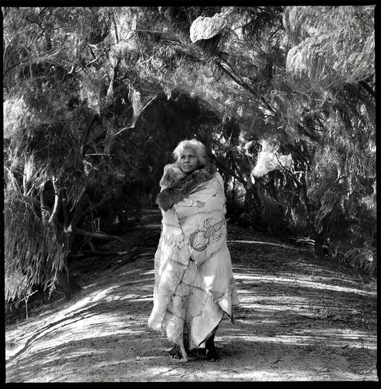 Black and white photograph of a woman standing on a dirt path with a possum skin cloak wrapped around her, encircled by a canopy of tree branches overhead.