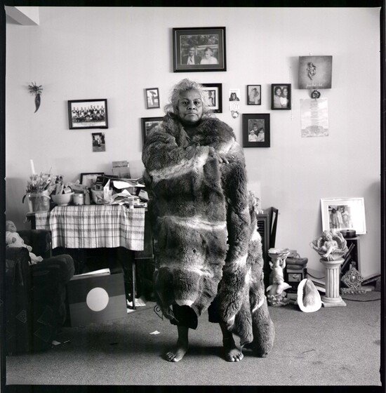 Black and white photograph of a woman standing in the centre of a lounge room, wrapped in a possum skin cloak. Behind her are many gramed photographs hanging on the wall, a table with a table cloth covered in papers and household items. A board painted with the Aboriginal flag rests against the table, and there are other household items on the carpeted floor. 