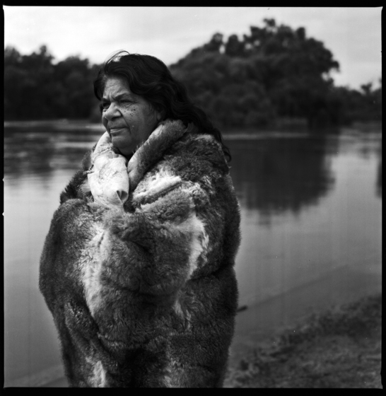 Black and white photograph of a woman standing on a river bank, with the river and trees across the bank behind, wrapped in a possum skin cloak.