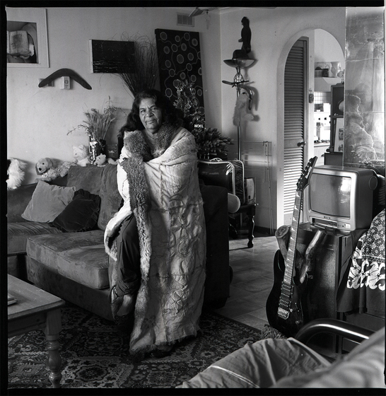 Black and white photograph of a woman sitting on the armrest of a couch in a living room, wrapped in a possum skin cloak. Beneath the couch is arug, with a small tv set and electric guitar to her right, with a boomerang and displayed artwork behind. A kitchen is seen through an archway behind.  