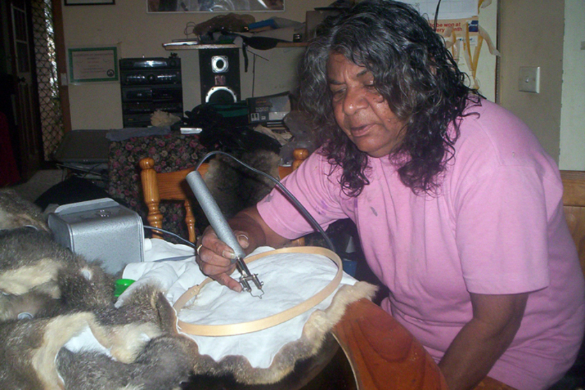 Woman in a pink tee shirt holding a soldering iron, to the inner side of a possum skin cloak laying on a table in front of her. 