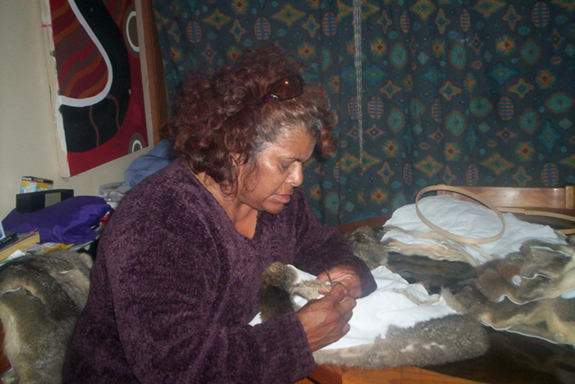 Woman seated at a table wearing a purple jumper, with her hands sewing the inner side of a possum skin cloak. There is a coloured green patterned curtain behind.