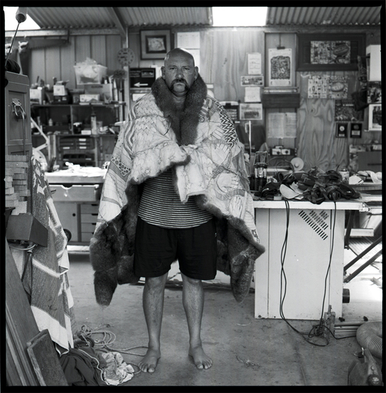 Black and white photograph of a man standing in a shed with a possum skin cloak hung over his shoulders. Behind are workbenches, and on the walls are shelves, tools and pictures.