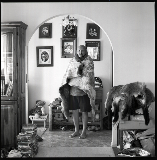 Black and white photograph of a man with a possum skin cloak wrapped around his shoulders, standing beneath an archway in a living room,. Behind are framed photographes, and a bench. Household goods can be seen surrounding.
