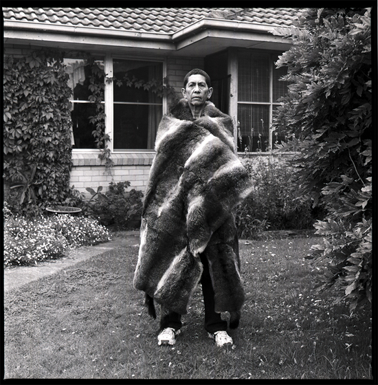 Black and white phorograph of a woman with a possum skin cloak wrapped around her shoulders standing on grass in front of a suburban house.