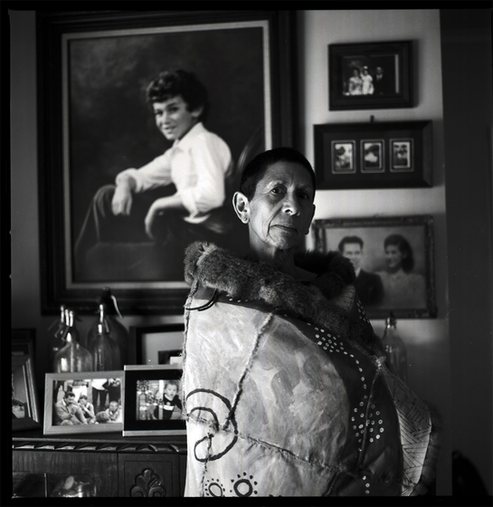 Black and white photograph  of a close up view of a woman with a possum skin cloak wrapped around her shoulders standing indoors, with a large painting of a young person on the wall behind, to the left ofthree smaller frames of photographs.of a woman's upper 