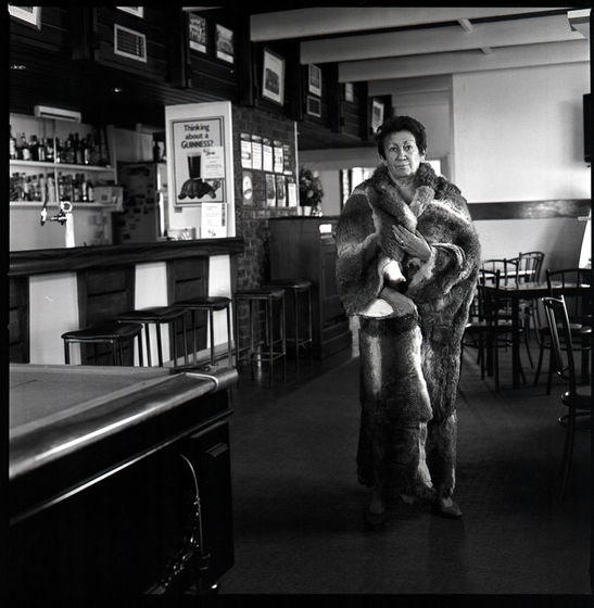 Black and white photograph of a woman with a possum skin cloak wrapped around her shoulders, standing on front of a hotel bar with stools in the background.