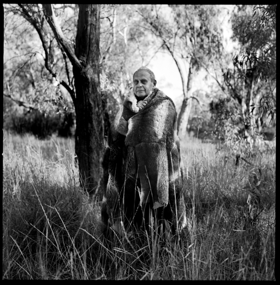 Black and white photograph of a man with a possum skin cloak wrapped around his shoulders, standing in tall grass in front of a gum tree.