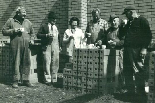 Five men and one woman share a meal on a pile of new bricks at a construction site