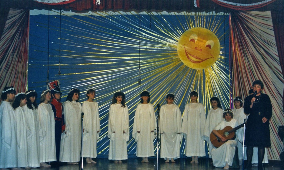 A group of 15 perform on a stage with a painted sunshine backdrop