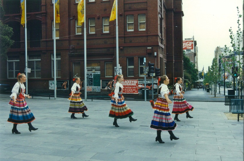 Six young women in traditional Polish costume dance in a large outdoor public square