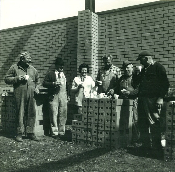 Five men and one woman share a meal on a pile of new bricks at a construction site
