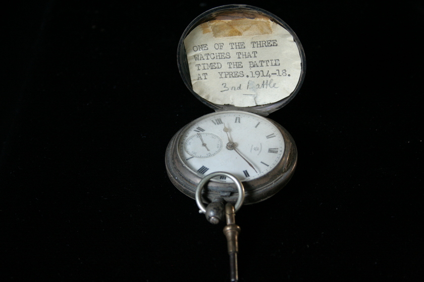 Open silver fob watch on a black background. The clock face is white with Roman numerals. The lid of the clock has a white card stuck inside with typed text 'ONE OF THE THREE WATCHES THAT TIMED THE BATTLE AT YPRES. 1914-18', and handwritten '3rd Battle'.