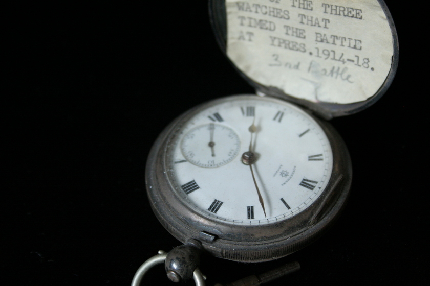 Close up view of open silver fob watch on a black background. The clock face is white with Roman numerals. The lid of the clock has a white card stuck inside with partial typed text seen reading '...THE THREE WATCHES THAT TIMED THE BATTLE AT YPRES. 1914-18', and handwritten '3rd Battle'.
