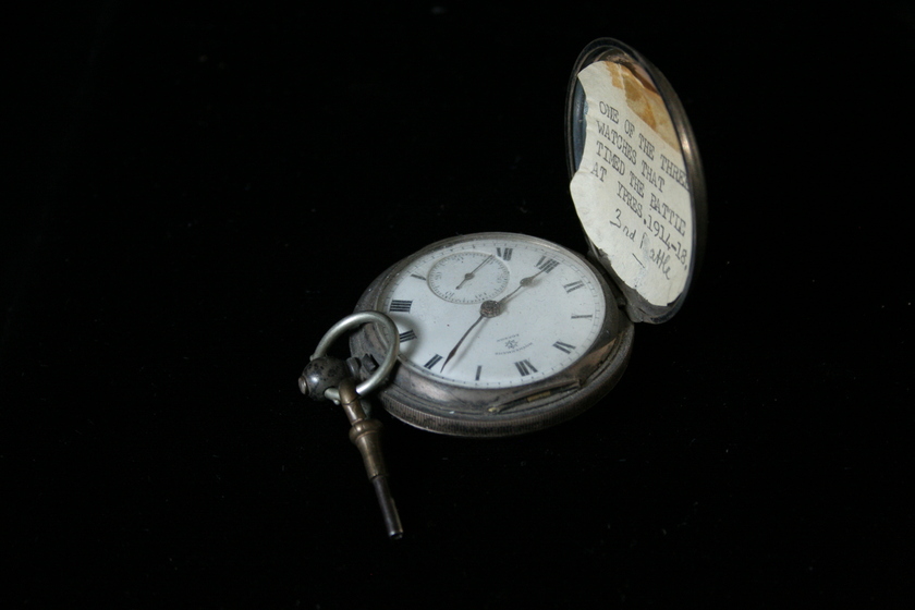 Side view of an open silver fob watch on a black background. The clock face is white with Roman numerals. The lid of the clock has a white card stuck inside with typed text.
