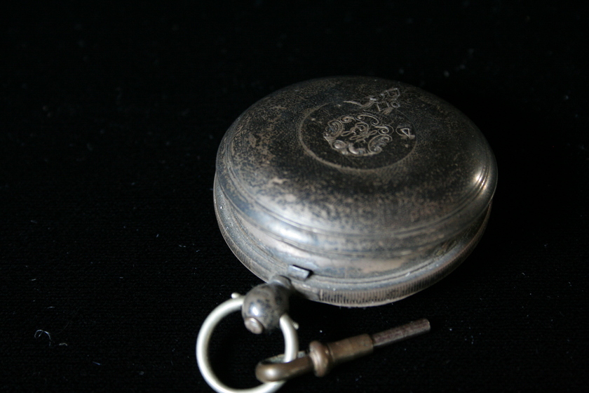 Closed silver fob watch on a black background, with intricate design on the lid. 