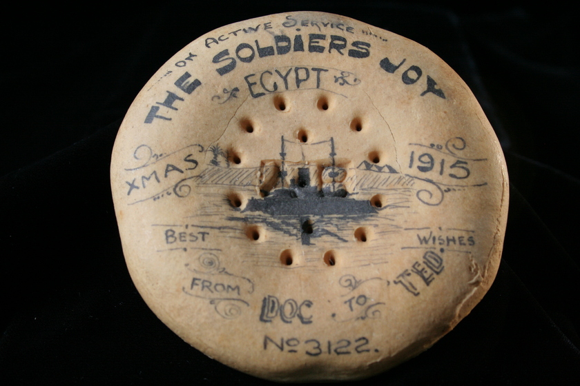 Round biscuit on a black background, covered in ink writing and illustrations. Text reads 'The Soldiers Joy, Egypt, Xmas 1915, Best Wishes from Doc to Ted". 