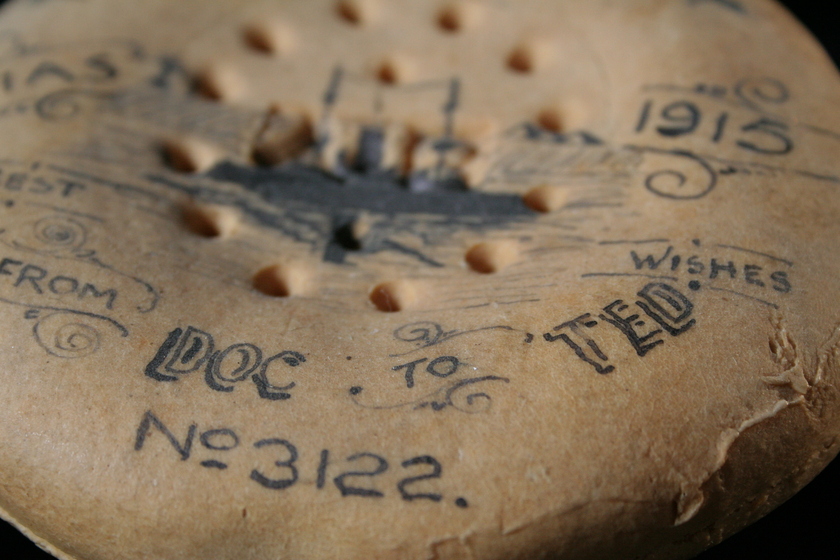 Close up view of part of a round biscuit on a black background, covered in ink writing and illustrations. Text seen reads 'Best Wishes from Doc to Ted". 