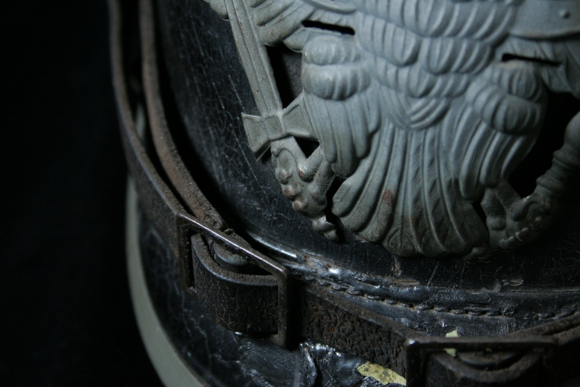 Close up view of feet of silver bird detail on black helmet, and leather strap and buckle below.
