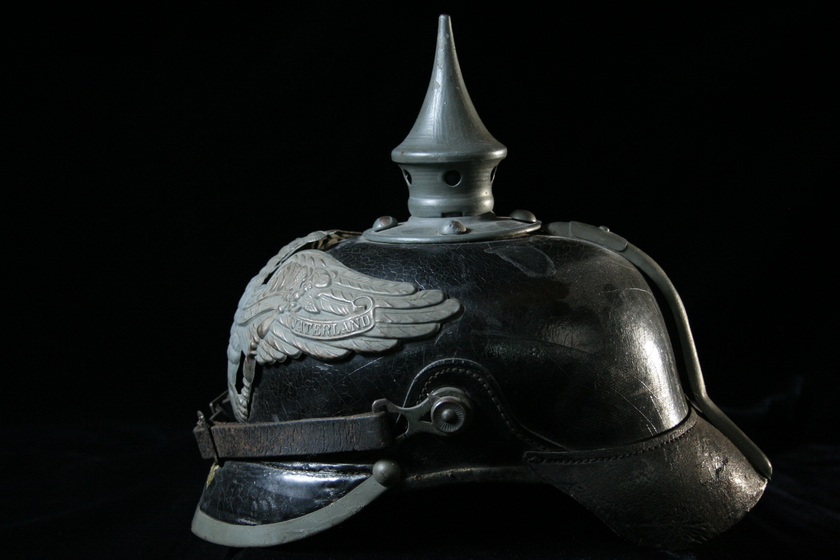 Side view of black helmet with silver spike on top, featuring silver open winged bird detail and a leather strap and buckle across the front.  