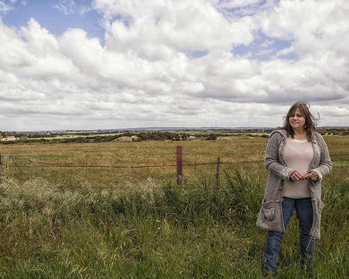 Woman standing in a grass field with a wire fence behind, and cloudy sky above.