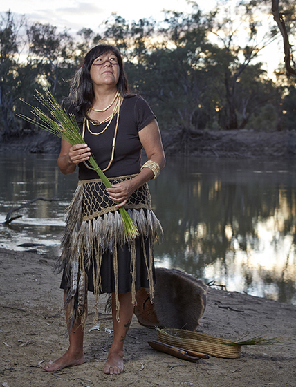 Woman in a black shirt, with a feather and twine skirt, holding a large bundle of green plant reeds, standing in front of a river.